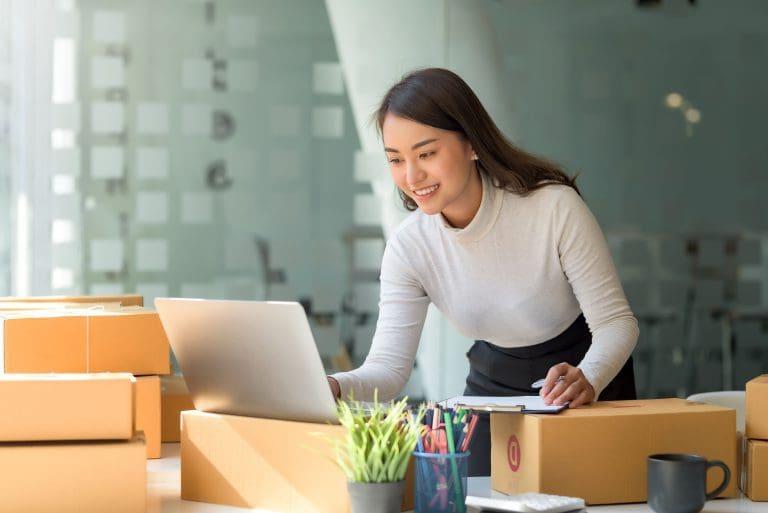 Happy young Asian business owner woman prepare parcel box and check online orders of product for deliver to customer on laptop computer. Shopping Online concept